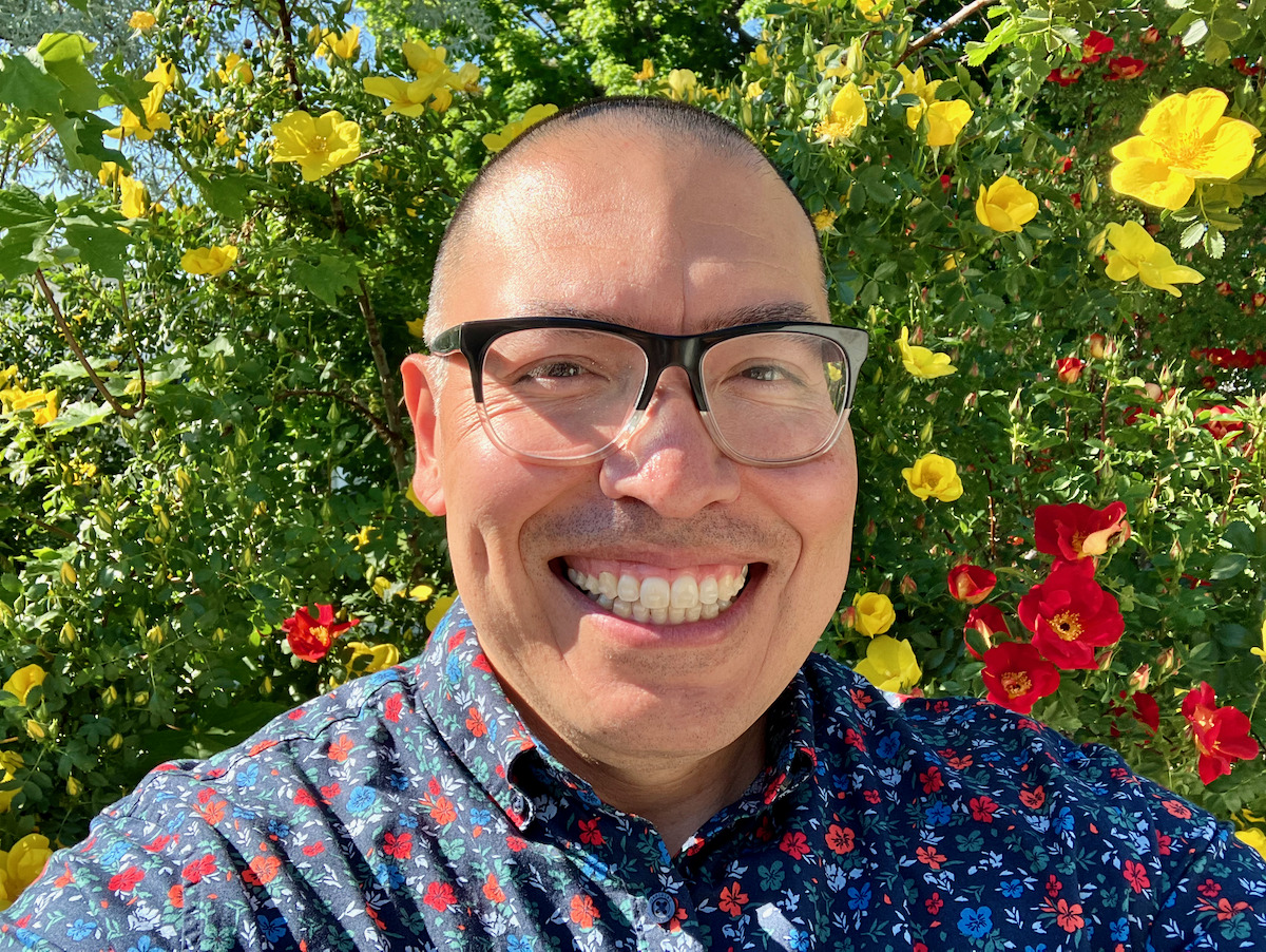 A picture of Ricardo Anguiano, wearing large
glasses and a flower-patterned collared shirt. The
background is a green hedge with yellow and red
flowers.